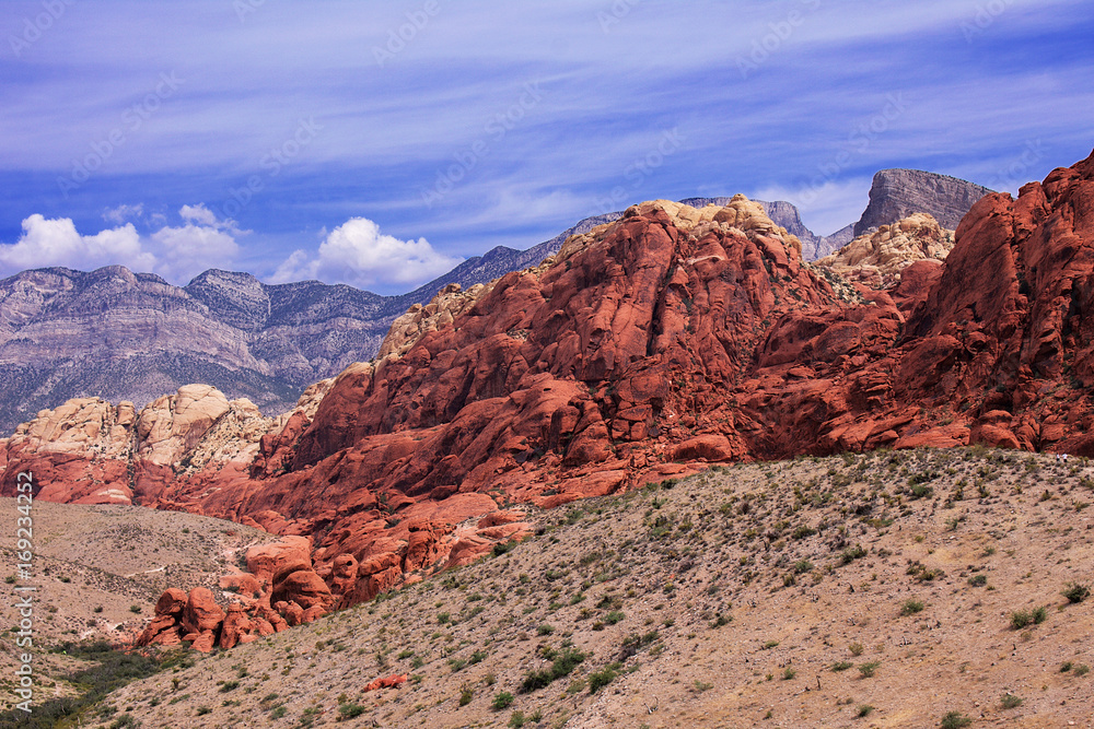 Mountain ranges in Red Rock, Nevada. The rocks are vivid red, orange and dark brown, and show signs of heavy erosion. The sky is blue and cloudy. The landscape is grand and dramatic and is reflective 