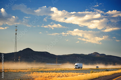 A white truck on a dusty freeway. In the background are dark brown hills and a dark blue sky with fluffy clouds. A mobile phone tower sits beside the road. In the foreground is light yellow foliage. T