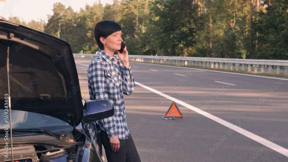 Young woman standing near broken car on the road. Caucasian brunette using smartphone calling for help. Attractive girl interrupted the trip because of a breakdown Young woman standing near broken car on the road. Caucasian brunette using smartphone calling for help. Attractive girl interrupted the trip because of a breakdown