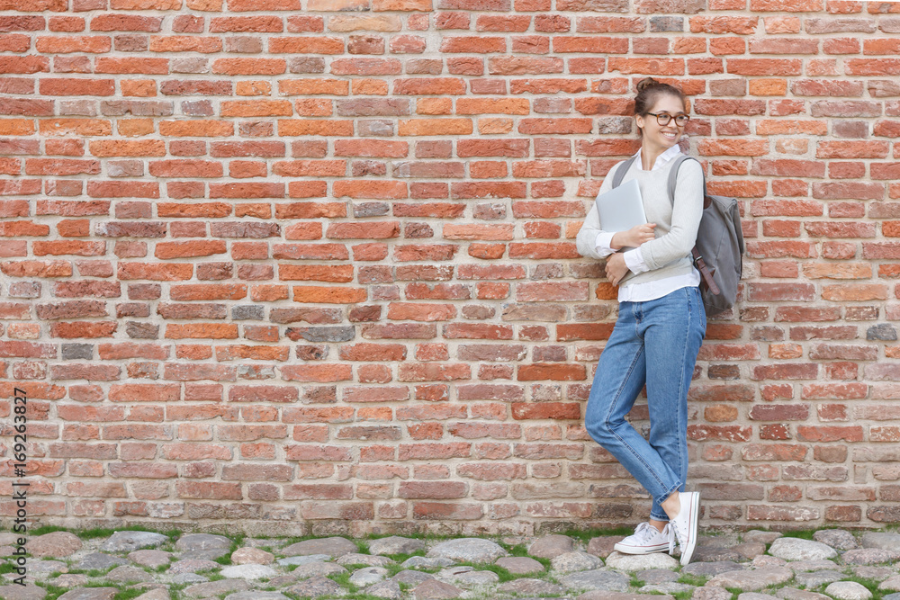 Full-height portrait of young beautiful woman leaning to red brick wall holding laptop in hands and turning rightwards as if awaiting for friend to come, looking happy and relaxed during her walk.