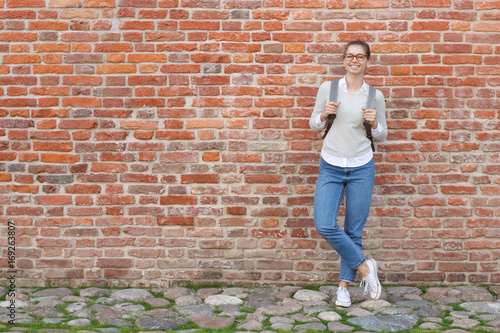 Full-height portrait of young European female standing against red brick wall, leaning to it with backpack, wearing casual clothes, looking happy and eager to continue journey alone or in company.