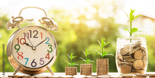 wooden house model and step of coins stacks with tree growing on top and clock alarm, nature background, money, saving and investment or family planning concept, over sun flare silhouette tone.