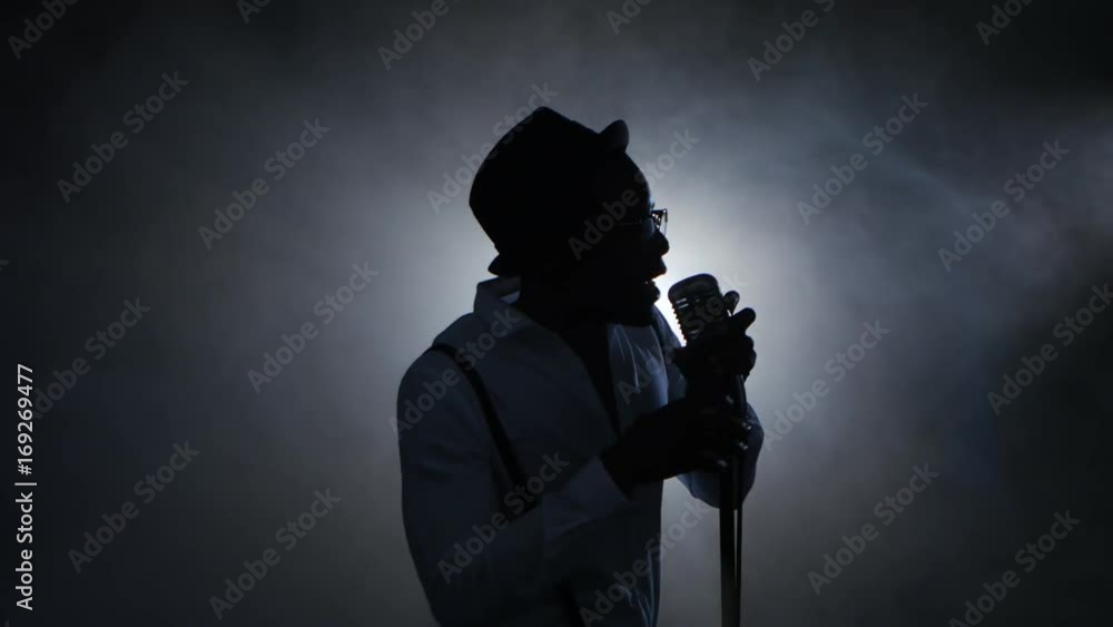 Man african american singer in the smoke and white light sings into a microphone and dance. Black background. Silhouette Man african american singer in the smoke and white light sings into a microphone and dance. Black background. Silhouette