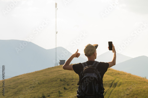 Male tourist on hiking trip happy to receive mobile network connection. Young man on vacation in mountains lifts thumb up and holds cell phone near the cell network tower