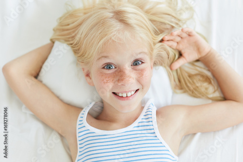 Aannemelijk Koe Yoghurt Cute little female child with blonde hair, blue eyes and freckled face,  smiling joyfully while relaxing on bed, lying on white pillow. Small  freckled girl in bed going to sleep. Children, lifestyle