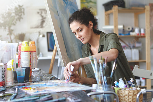 Picture of serious concentrated young Caucasian female artist sitting at desk with painting accessories, holding tube of oil paint, mixing colors on palette; unfinished painting on canvas near her photo