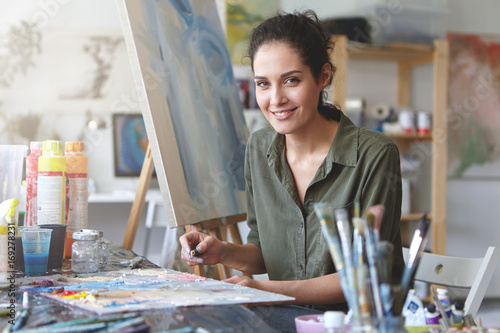 Portrait of young talented female artist making sketches with brightful oils, drawing on easel, having pleasant smile. Smiling painter being busy with her work at workshop. Art, creativeness concept photo