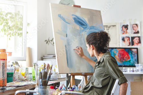 Art, creativity, hobby, job and creative occupation concept. Rear view of busy female artist sitting on chair in front of easel, painting with fingers, using white and blue oil or acrylic paint photo