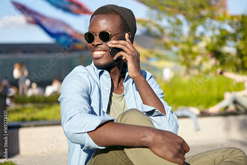 Positive young male with dark skin, smiling broadly while having conversation with his best friend, speaking over smart phone while resting outdoors. People, communication, technology concept