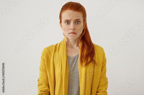 Scared red-haired woman with pony tail, stairing at camera, curving her lips being shocked to realize something horrorful. Shocked young female in yellow clothes isolated over white background photo
