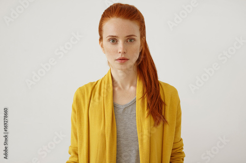 Horizontal portrait of beautiful young female lady with freckles ang ginger pony tail, wearing yellow clothes, having full lips and green shining eyes, looking directly into camera with confidence