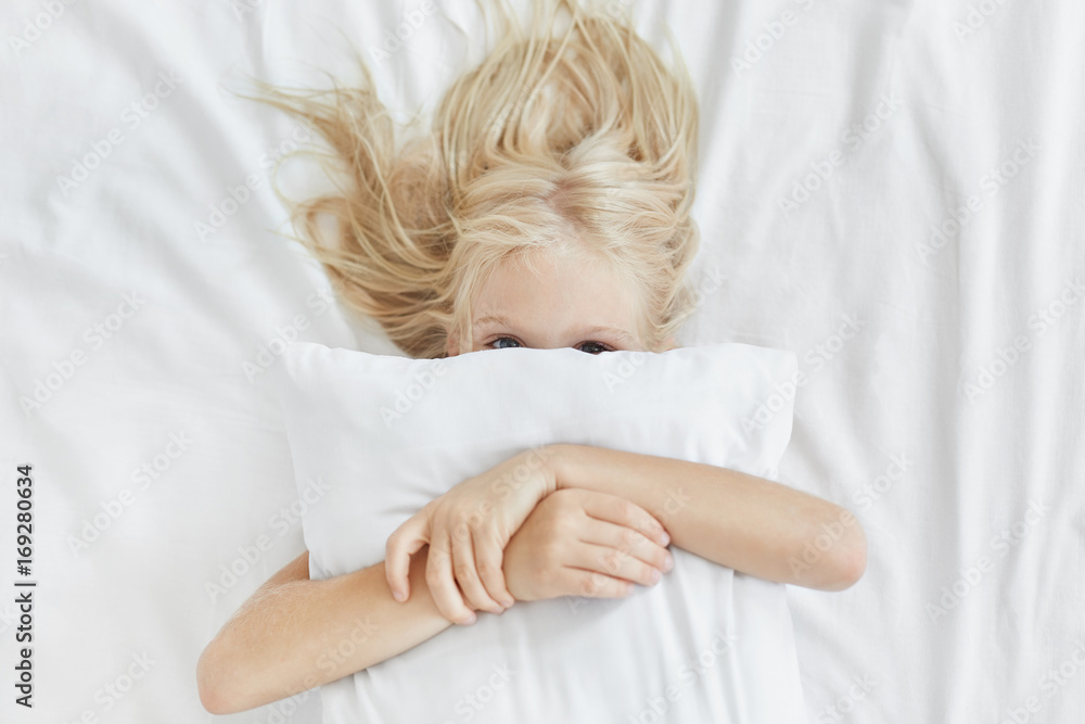 Sweet cute girl with light long hair, hiding behind white pillow, embracing it while lying in bed, having fun in morning. Playful child hugging pillow covering face with it. Health, sleep and children Sweet cute girl with light long hair, hiding behind white pillow, embracing it while lying in bed, having fun in morning. Playful child hugging pillow covering face with it. Health, sleep and children