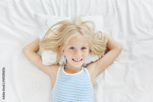 Schilderij op canvas Lovely blue eyed female child with freckles, lying on white pillow, keeping hands behind it, smiling pleasantly into camera, being glad to see her parents in her bedroom