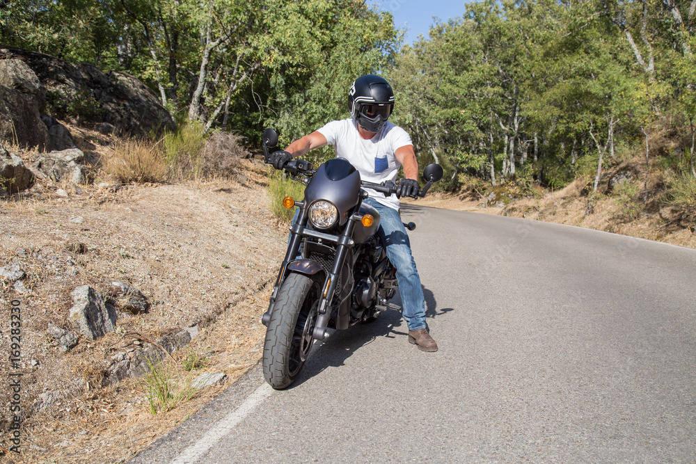 Side view of man sitting on parked custom motorcycle on road in mountains. Side view of man sitting on parked custom motorcycle on road in mountains.