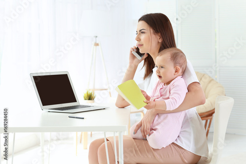 Young mother holding baby while talking on phone in home office