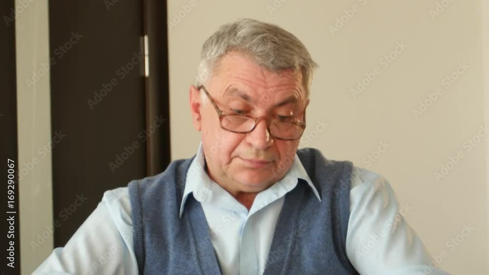 Gray haired man unravels crossword puzzles, he has poor eyesight and he is wearing glasses Gray haired man unravels crossword puzzles, he has poor eyesight and he is wearing glasses