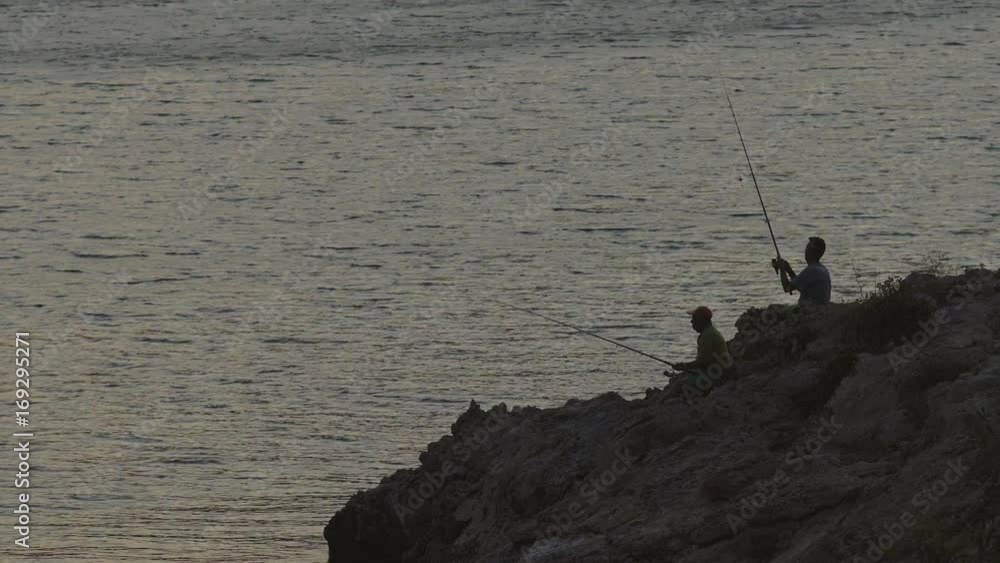 Two Unidentifiable Adult Men In Silhouette Fishing At Dusk By The Sea On A Rocky Shore In Summer. Two Unidentifiable Adult Men In Silhouette Fishing At Dusk By The Sea On A Rocky Shore In Summer.