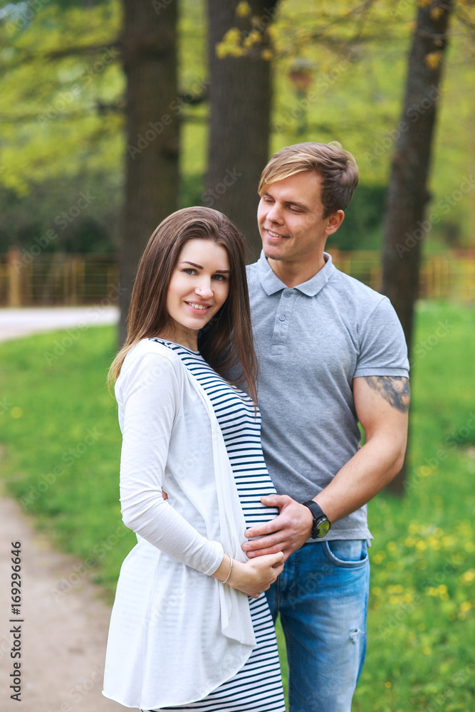 Handsome man and his beautiful pregnant wife are hugging and smiling while standing in park Handsome man and his beautiful pregnant wife are hugging and smiling while standing in park