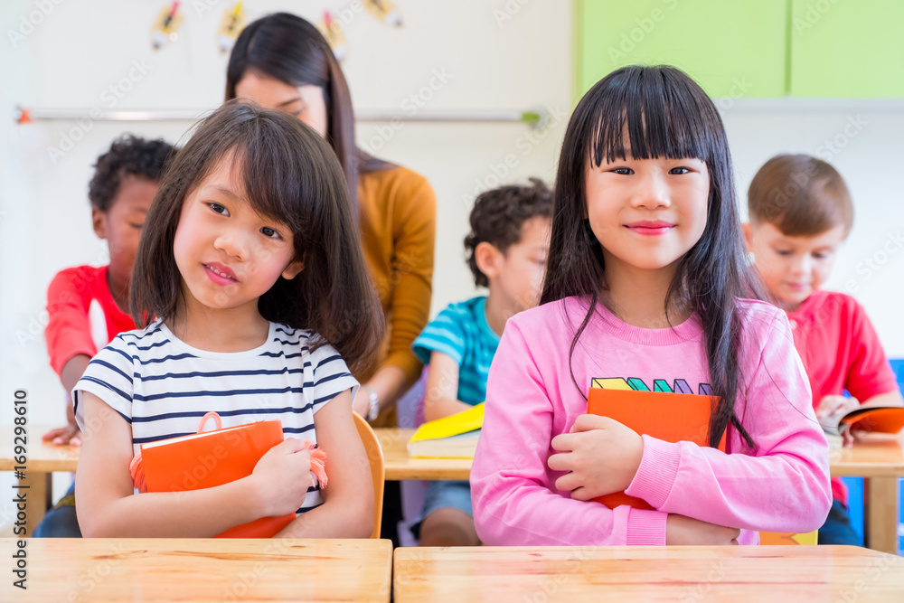 Two Asian girl kid hugging book and smiling in classroom and while teacher teach friends beside them,kindergarten education Two Asian girl kid hugging book and smiling in classroom and while teacher teach friends beside them,kindergarten education
