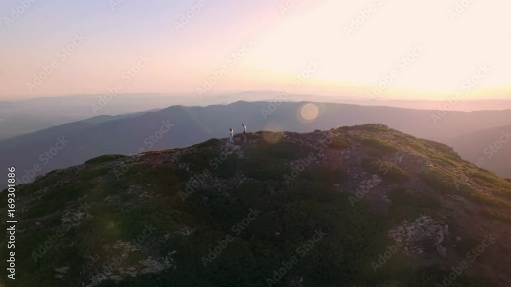 Beautiful summertime lifestyle drone video of attractive young hipster millennial couple descending mountain ridge, with purple pink sunset or sunrise bursting in colors behind them Beautiful summertime lifestyle drone video of attractive young hipster millennial couple descending mountain ridge, with purple pink sunset or sunrise bursting in colors behind them