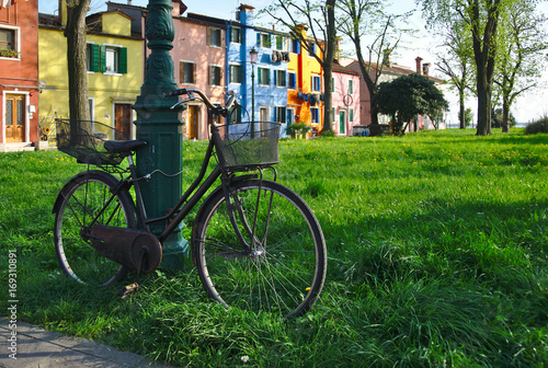 Old black city bicycle with basket chained to a pole in grass in front of colorful houses in Burano island in summer