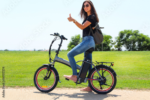 A smiling woman with electric bike in the park Makes a like with her hand