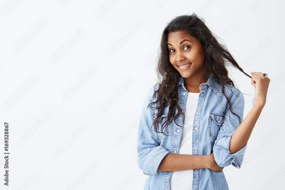 Studio shot of cute dark-skinned woman playing with her long wavy hair, glowing face and gentle smile rejoicing her beauty. Cheerful woman dressed casually having pleased and flirting expression Studio shot of cute dark-skinned woman playing with her long wavy hair, glowing face and gentle smile rejoicing her beauty. Cheerful woman dressed casually having pleased and flirting expression