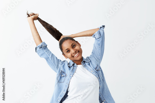 Indoor shot of beautiful afro-american girl with long black hair gathered in ponytail dressed casually posing indoors, adjusting her hairstyle before going out. People and emotions concept. photo