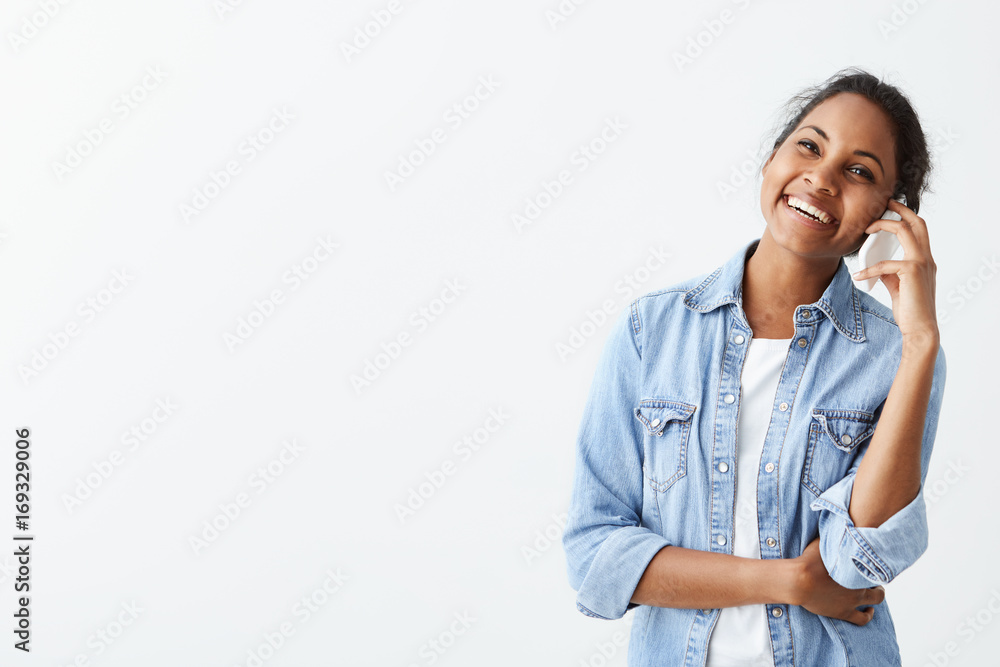 Happy young Afro-american woman in blue shirt with dark hair discussing her meeting on smartphone looking aside with charming appealing smile. Happy young Afro-american woman in blue shirt with dark hair discussing her meeting on smartphone looking aside with charming appealing smile.