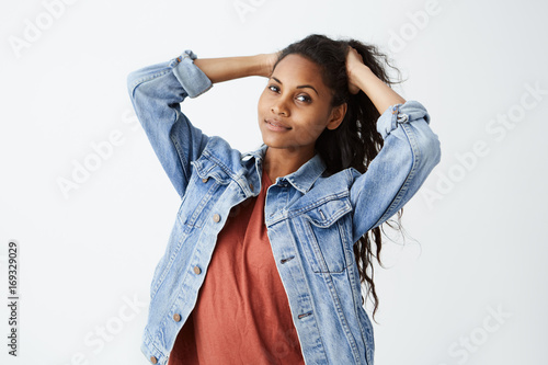 Fashionable young dark-skinned woman wearing denim jacket and red casual t-shirt tying her beautiful dak wavy hair while posing. Horizontal studio shot photo