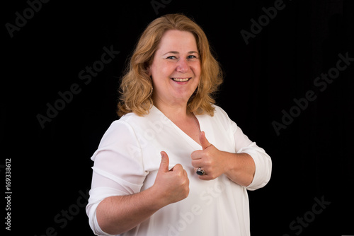 Portrait of a attractive european light overweighted red haired female in white shirt showing thumbs up in front of black backgrouind