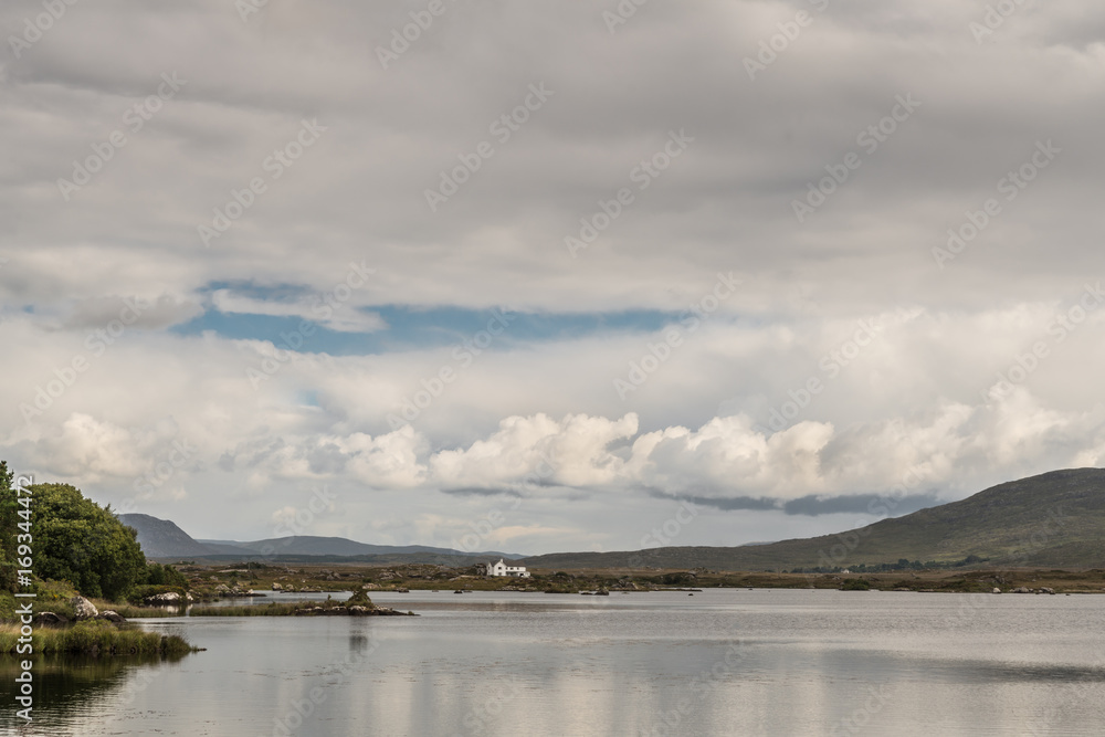 Connemara County, Ireland - August 4, 2017: Wide view of Furnace Lake under a gray sky with one blue patch and pronounced low clouds. Green hills and a white house on the horizon.