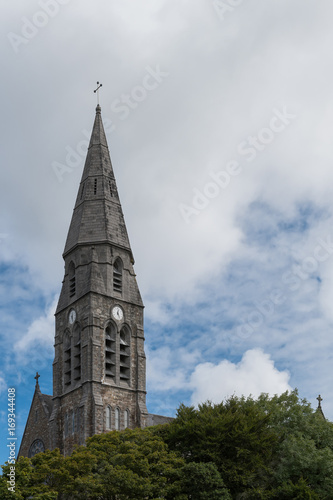 Clifden, Ireland - August 4, 2017: the gray spire of the local Catholic church tower against white clouds in blue sky. Some green of trees at bottom.