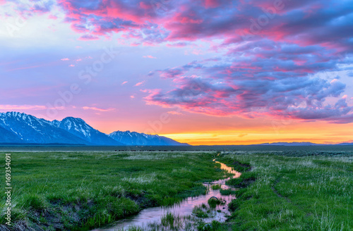 Fototapeta Sunset Mountain Meadow - Colorful spring sunset at a green mountain field with a winding stream near Mormon Row historic district in Grand Teton National Park, Wyoming, USA