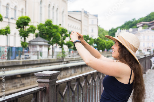 Woman taking pictures with smartphone. Stylish summer traveler woman in hat with camera outdoors in european city  old town Karlovy Vary in the background  Czech Republic  Europe