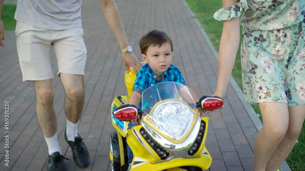 Beautiful family is walking in the park while their little cute son is riding adorable yeallow motorcycle Beautiful family is walking in the park while their little cute son is riding adorable yeallow motorcycle