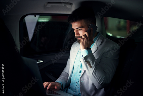 Elegant modern businessman with laptop on the back seat talking on a mobile. © dusanpetkovic1