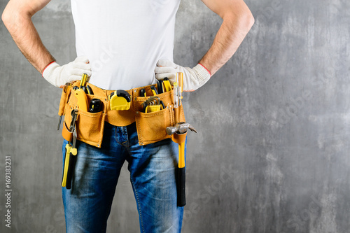 Photography unknown handyman with hands on waist and tool belt with construction tools against grey background with copyspace for text