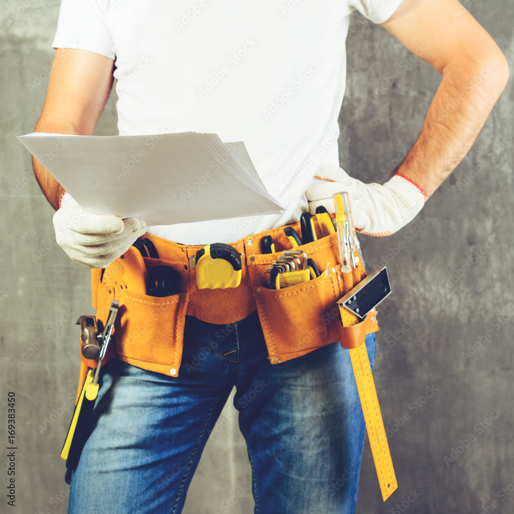 unknown handyman with hand on waist and tool belt with construction tools holding the project plane against grey background, toned image. DIY tools and manual work concept unknown handyman with hand on waist and tool belt with construction tools holding the project plane against grey background, toned image. DIY tools and manual work concept