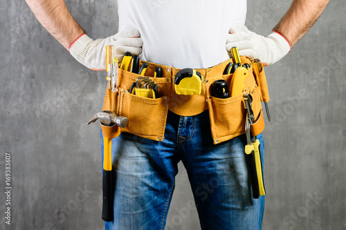 unknown handyman with hands on waist and tool belt with construction tools against grey background. DIY tools and manual work concept photo