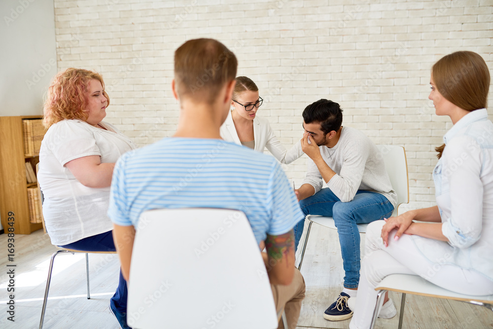 Fototapeta premium Portrait of young man crying in support group sharing problems with female psychiatrist