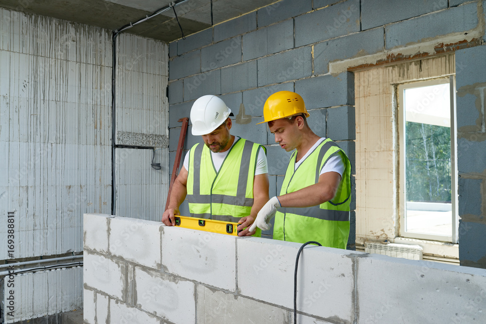 Portrait of two construction workers wearing hardhats and reflective vests checking concrete block wall using level Portrait of two construction workers wearing hardhats and reflective vests checking concrete block wall using level