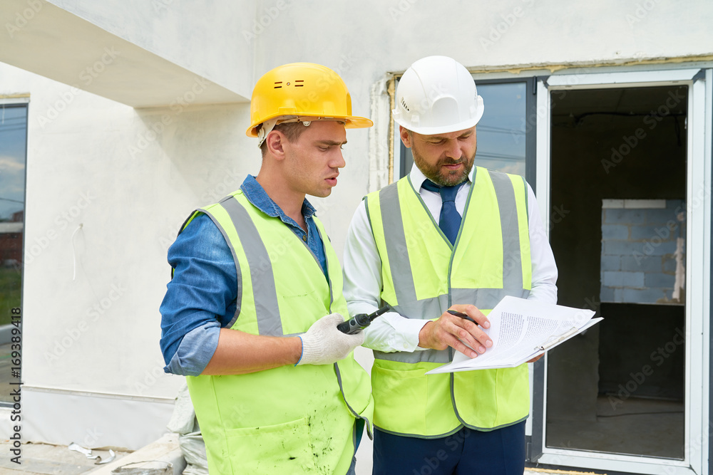 Portrait of foreman and supervisor discussing construction documents on site, both wearing hardhats and reflective vests Portrait of foreman and supervisor discussing construction documents on site, both wearing hardhats and reflective vests