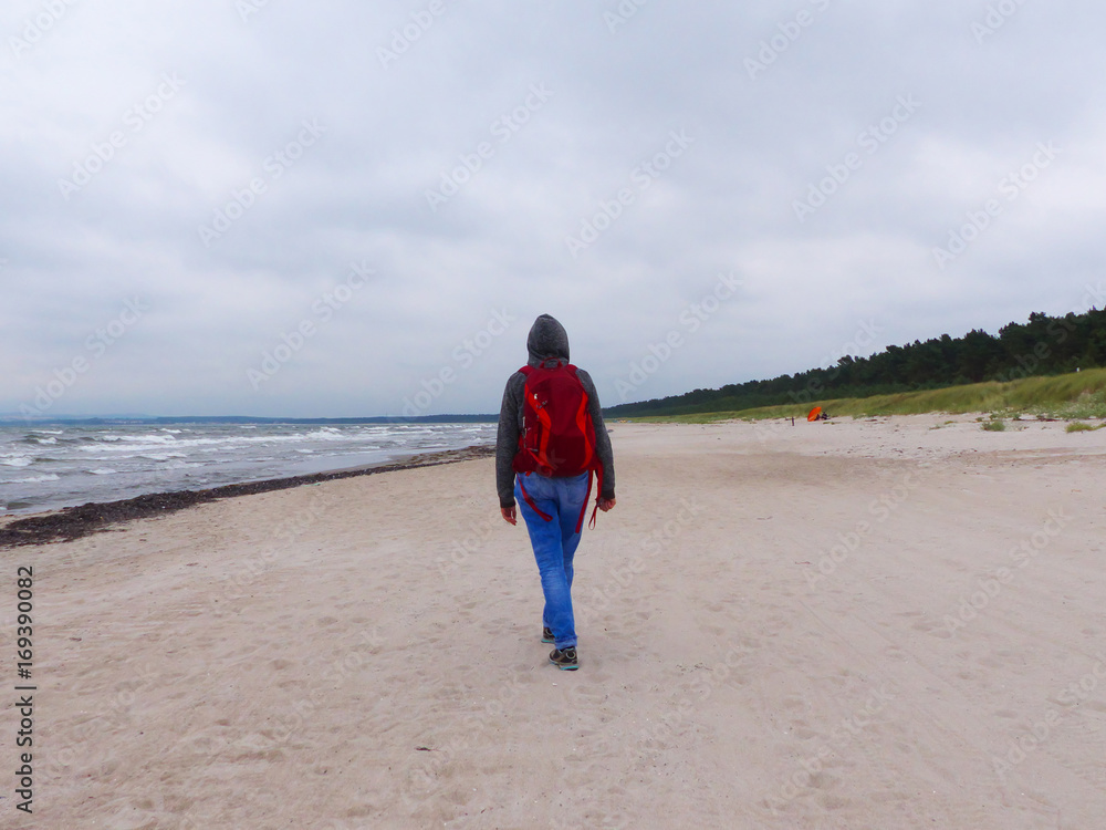 Spaziergang am Strand bei grauem Wetter, Frau im Zentrum mit rotem Rucksack