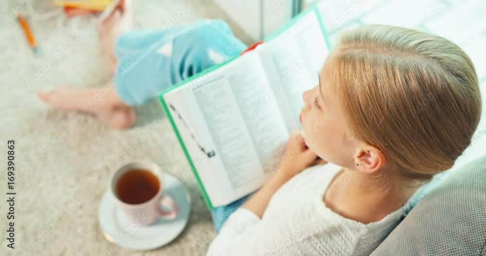 Girl reading book sitting on the floor near window and smiling at camera. Top view Girl reading book sitting on the floor near window and smiling at camera. Top view