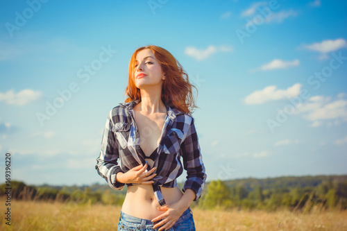 Red-haired attractive American style girl, checkered shirt and black bra and shorts standing in a meadow at sunset enjoying the rays of the sun