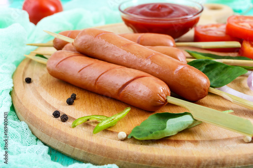 Juicy sausages on skewers with tomatoes and tomato sauce on a round board on a bright background. Top view.