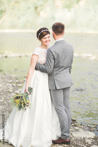 Handsome romantic groom and beautiful bride posing near river in scenic mountains