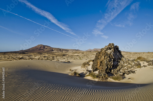 Felsbrocken aus Lavagestein in einer Sandmulde einer D  nenlandschaft mit flachen Bergen im Hintergrund.  
