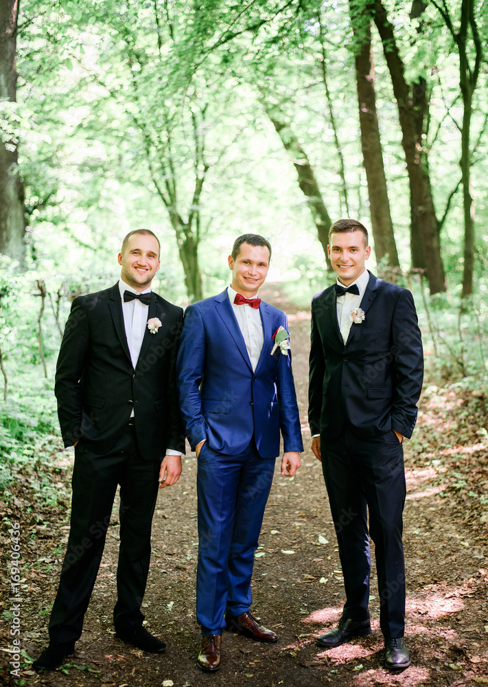 Groom in bright blue suit and gromsmen in black tuxedo stand on path in the forest Groom in bright blue suit and gromsmen in black tuxedo stand on path in the forest